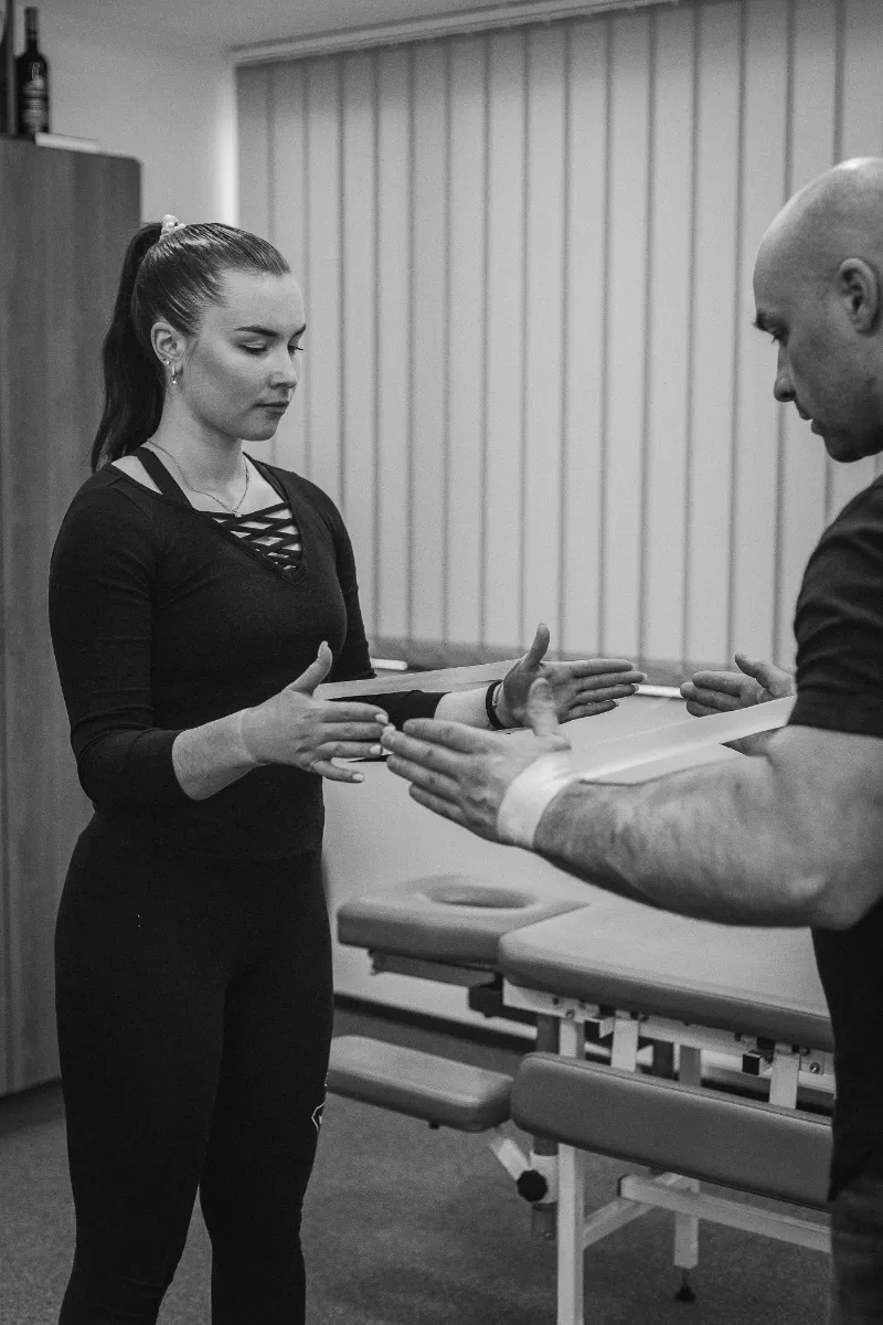 Mustvalge foto: naine seisab vasakul, osteopaat Ruslan Mursalov paremal, mõlemad venitavad käte vahel kummilinti.